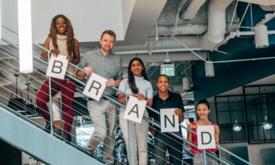 A diverse team standing on an office staircase holding letters that spell "BRAND," representing branding, public relations, and teamwork.