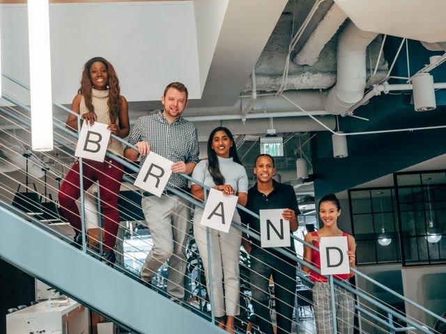 A diverse team standing on an office staircase holding letters that spell "BRAND," representing branding, public relations, and teamwork.